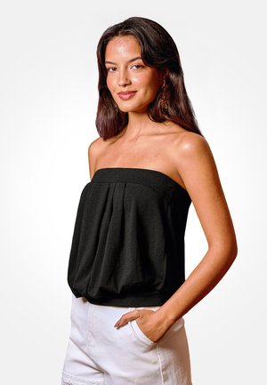 Jonge vrouw met donker haar draagt een zwart strapless topje en witte shorts, staand met één hand in de zak tegen een effen achtergrond.