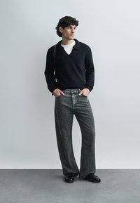 Pull noir en maille avec col en V, associé à un jean en denim gris à coupe ample. Des chaussures noires complètent la tenue, le tout sur un fond neutre.