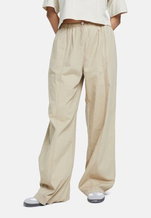 Personne portant un pantalon large beige avec une taille élastique et des baskets blanches, debout devant un fond uni.