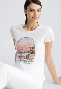 T-shirt blanc avec un imprimé coloré représentant une scène de plage comprenant deux chaises, un parasol, et le texte "Vibes d'été". Texture de coton lisse.