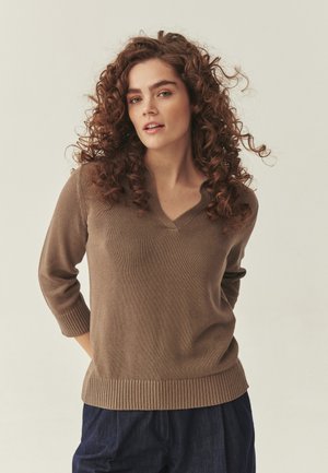 Młoda kobieta o kręconych włosach, ubrana w brązowy sweter z dekoltem w serek i ciemnoniebieskie spodnie, pozuje z rękami za plecami na jednolitym tle.