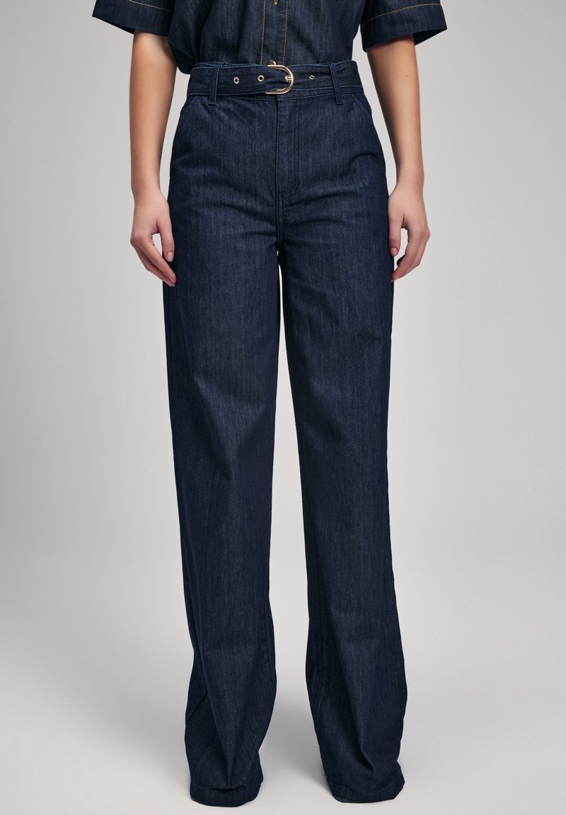 Femme portant un jean large taille haute bleu foncé avec une ceinture assortie à boucle dorée, debout devant un fond uni.