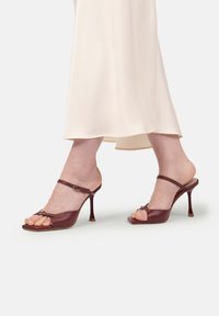 Picioare încălțate cu sandale cu toc înalt, strânse, de culoare burgundă, asortate cu o fustă bej deschis, fluidă, pe un fundal simplu.