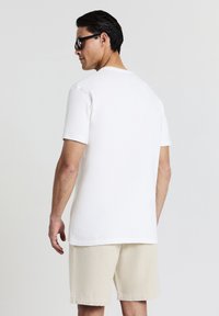 T-shirt bianco a maniche corte con finitura testurizzata, abbinato a pantaloni corti beige. Il modello indossa occhiali da sole neri, visto da dietro.