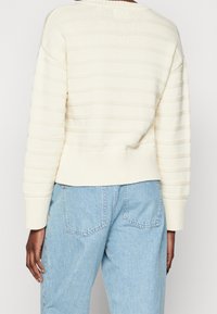 Suéter de punto a rayas crema con ribetes en el dobladillo y los puños, que presenta un ajuste relajado. Combinado con jeans de mezclilla de talle alto en azul claro.