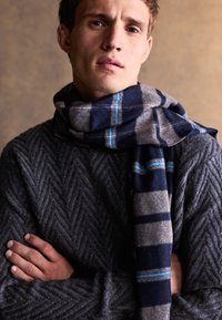 Šedý svetr s texturou a rybí kostí. Vlnená šála v námořnické modři, šedé a modré kárované vzory je volně přehozena kolem krku. Ruce zkřížené.