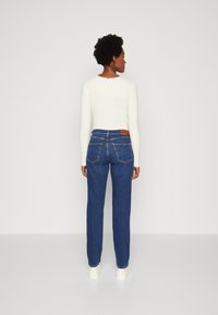 Jeans in denim blu presentano un classico design a gamba dritta, cinque tasche e una patch in pelle marrone sul retro. Visibile una maglietta a maniche lunghe di colore crema.