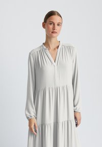 Robe gris clair à manches longues avec col en V, corsage froncé et jupe à volants. Confectionnée en tissu lisse avec un léger éclat et des poignets élastiques.