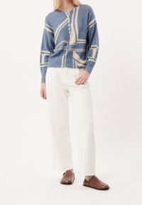Femme portant un pull boutonné à motifs bleus, un pantalon large blanc, des chaussettes beiges et des chaussures en cuir marron sans lacets, sur un fond uni.