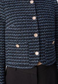 Veste en tweed noire et bleue coupée mettant en avant un motif texturé, des boutons dorés et un ourlet arrondi. Associée à un pantalon noir.