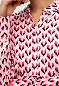 Camicia bianca con un motivo a zigzag nei colori rosa, nero e bianco; presenta bottoni e maniche lunghe con dettagli sui polsini. Tessuto morbido.