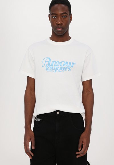 White cotton t-shirt with light blue graphic text "Amour Toujours" and "In Paris Summer 25" on the front; paired with black pants.