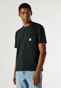 Μαύρο βαμβακερό t-shirt με στρογγυλή λαιμόκοψη, κοντά μανίκια και τσέπη στο αριστερό μέρος του στήθους που διαθέτει ένα μικρό λογότυπο. Φορεμένο με ανοιχτόχρωμο μπλε τζιν παντελόνι.