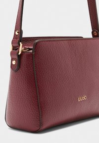 Borsa in pelle bordeaux con finitura texture, accenti in hardware dorato e base piatta. Logo del marchio sottilmente impressa sulla parte anteriore.