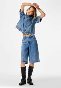 Moteris ištiesia rankas virš galvos, dėvi trumpomis rankovėmis džinsinę marškinių palaidinę, kelio ilgio džinsinius šortus ir juodus kelio aukščio batus ant baltos fono.