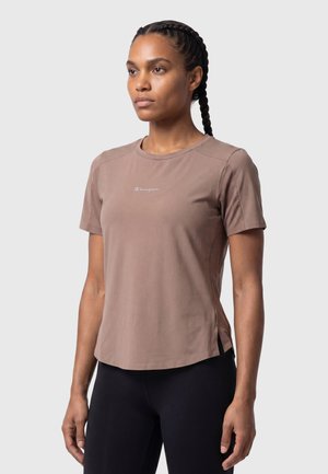 Vrouw met ingevlochten haar, gekleed in een lichtbruine Champion T-shirt met korte mouwen en een zwarte broek, staand voor een effen grijze achtergrond.
