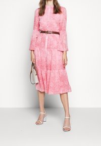 Robe rose à motif avec manches longues, design en volants et taille froncée, complétée par une ceinture marron et des sandales blanches à talons hauts.