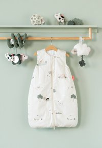 Saco de dormir blanco para bebé con dibujos de animales y árboles colgado en una percha de madera, rodeado de juguetes suaves para bebé en un estante verde de pared.