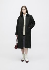 Femeie în picioare, purtând o haină lungă neagră, un pulover bej, o cămașă albă, o fustă gri, ținând o geantă burgundă, șosete albe și pantofi burgunzi.