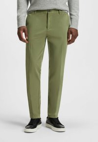 Green trousers with a smooth texture, featuring a button closure and side pockets, paired with black sneakers and a light gray sweater.