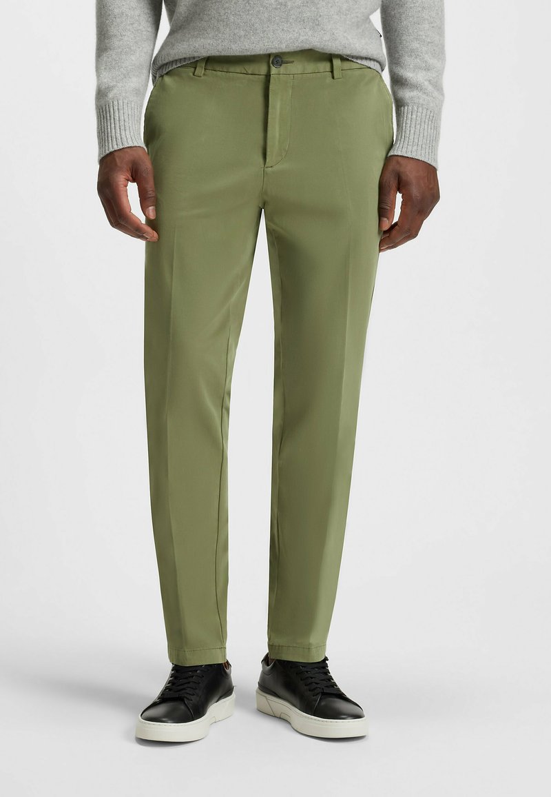 Green trousers with a smooth texture, featuring a button closure and side pockets, paired with black sneakers and a light gray sweater.