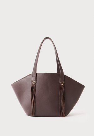 Sac fourre-tout en cuir marron avec deux poignées et des pampilles décoratives pendantes à chaque poignée sur un fond clair uni.