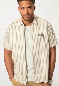 Camicetta beige a maniche corte con righe verticali blu, zip frontale e grafica "EAST COAST" sul lato sinistro del petto. Abbinata a una maglietta bianca.