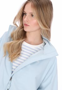 Világoskék esőkabát cipzáros lezárással és magas gallérral, fehér és világoskék csíkos ing fölött, sima textúrájú.