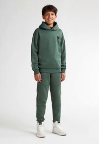 Completo con felpa verde con cappuccio e pantaloni della tuta caratterizzato da un logo, texture morbida e polsini a coste. Indossato con sneakers bianche per un look completo.