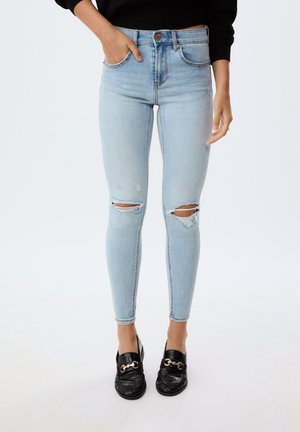 Jeans skinny bleu clair avec des déchirures aux genoux, design à cinq poches et fermeture à bouton, associés à des mocassins noirs avec des accents dorés.