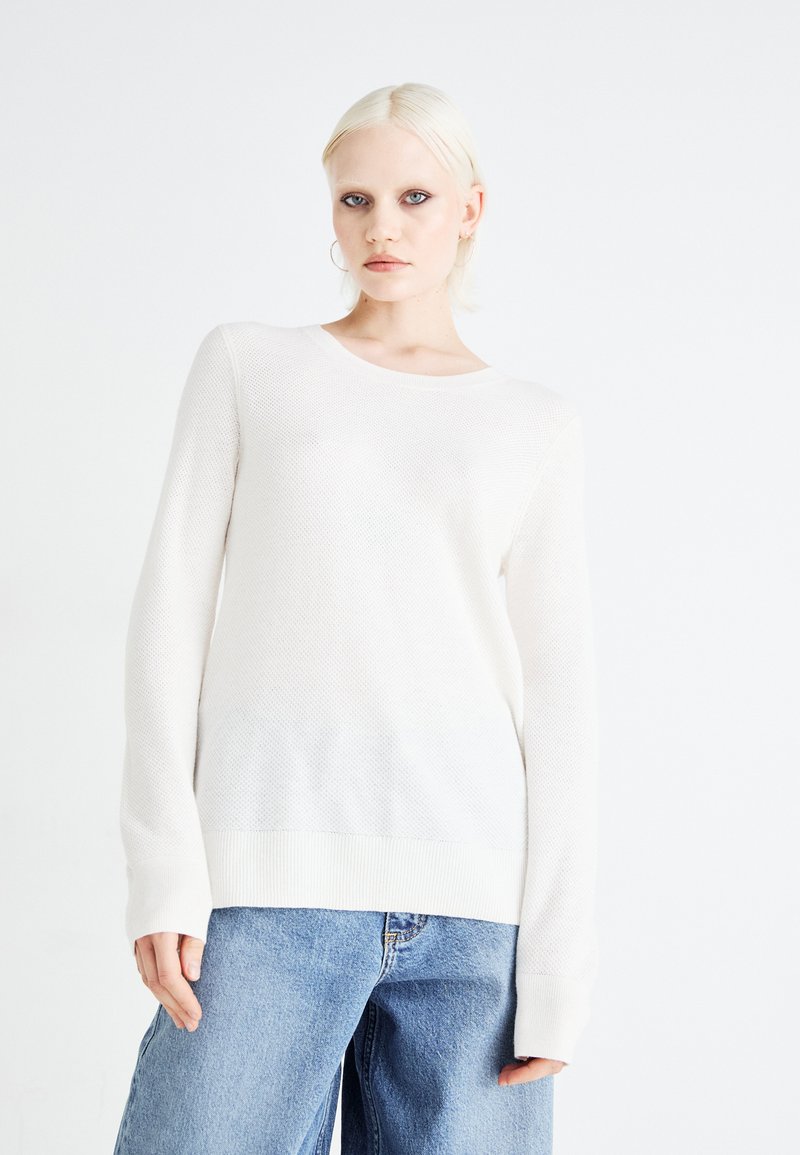 Pull en tricot beige clair avec un col rond et des manches longues. Présente un motif texturé et un ourlet côtelé. Associé à un jean ample bleu.