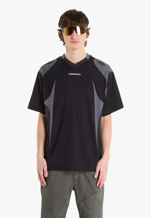 Giovane uomo con maglietta sportiva Umbro nera e grigia, occhiali da sole avvolgenti riflettenti e pantaloni grigi, in piedi contro uno sfondo bianco.