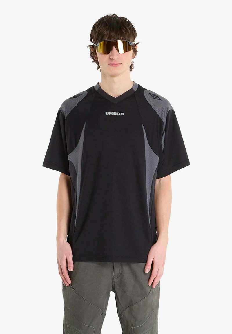 Giovane uomo con maglietta sportiva Umbro nera e grigia, occhiali da sole avvolgenti riflettenti e pantaloni grigi, in piedi contro uno sfondo bianco.