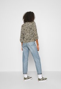 Blusa de estampado negro y marrón con mangas tres cuartos, combinada con jeans de color azul claro y zapatillas verdes oliva, que llevan calcetines blancos.