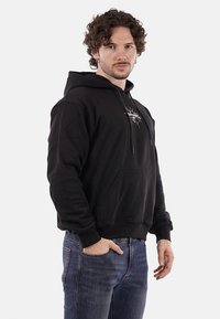 Felpa nera con cappuccio caratterizzata da una tasca a marsupio frontale, polsini e orlo a coste, e una stampa grafica sul petto. Indossata con jeans blu.