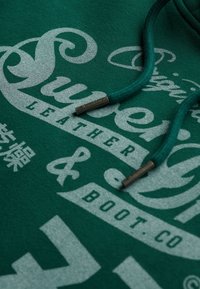 Hoodie turquoise avec un grand texte graphique argenté affichant "Super" et "Leather & Boot Co." Le texte comprend des accents et des cordons de serrage avec embouts métalliques.