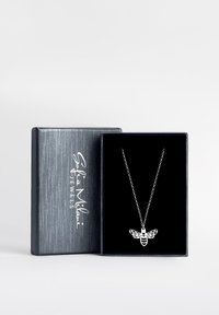 Silverhalsband med en bi-pendant med intrikata urklippsdetaljer, visad i en mörk sammetstoppad svart låda. Lådan har en texturerad grå utsida.