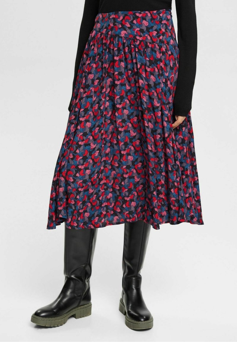 Femme portant une jupe mi-longue à imprimé abstrait rouge, rose et bleu, associée à des bottes longues noires et un haut à manches longues noir.