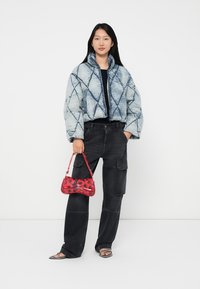 Vrouw in een blauwe gewatteerde jas, zwarte cargobroeken, sandalen, en met een rode bloemenhandtas tegen een witte achtergrond.