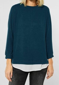 Kvinna som bär en mörk teal långärmad tröja över en vit skjorta, tillsammans med svarta jeans, visad från axlarna till mitten av låren.