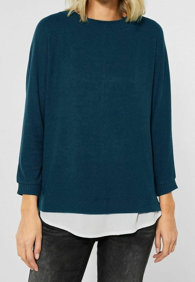 Kvinna som bär en mörk teal långärmad tröja över en vit skjorta, tillsammans med svarta jeans, visad från axlarna till mitten av låren.