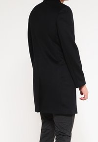 Cappotto di lana nero con maniche lunghe, taglio dritto e colletto. Presenta una texture liscia e non ha hardware o decorazioni visibili.