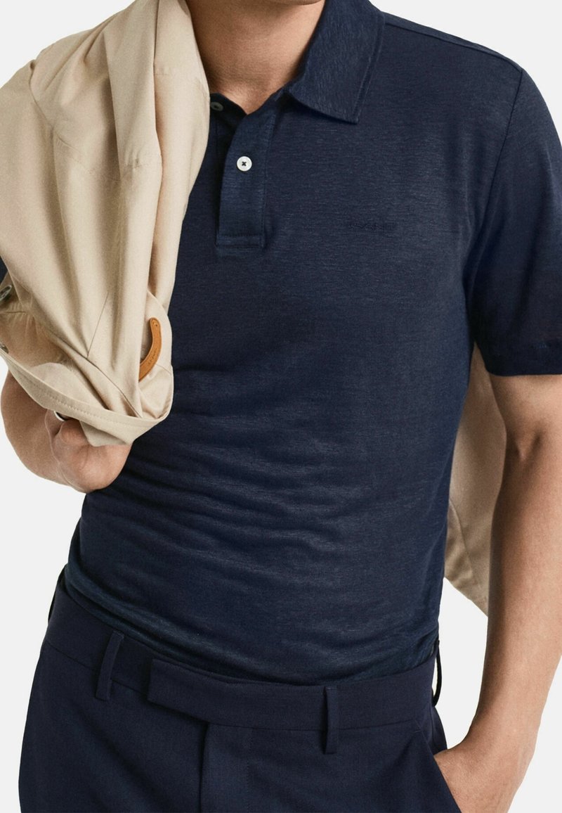 Polo bleu marine en tissu texturé, avec une patte à deux boutons et une veste beige clair bien pliée posée sur l'épaule.