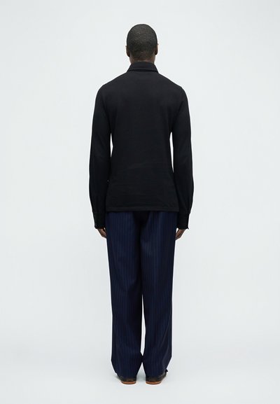 Άνδρας στέκεται με την πλάτη γυρισμένη φορώντας μαύρο μακρυμάνικο πουκάμισο, μπλε σκούρο φαρδύ παντελόνι με ρίγες και μαύρα παπούτσια σε απλό φόντο.