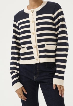 Marineblauw-witte horizontaal gestreepte cardigan met gouden knopen en steekzakken, gedragen met donkerblauwe jeans.