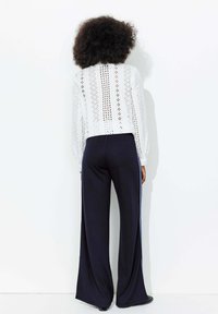 Blouse blanche à manches longues avec des motifs découpés complexes, associée à un pantalon large bleu marine avec une bande latérale.