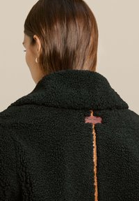 Giacca in pile nera con texture, dotata di colletto a scialle, caratterizzata da un'etichetta in pelle e una cucitura a contrasto lungo la schiena.