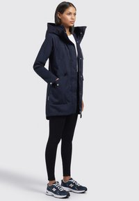 Granatowa parka z kapturem, z zamkiem błyskawicznym i dwoma przednimi kieszeniami, noszona na czarne legginsy i buty sportowe. Gładki materiał, casualowy design.
