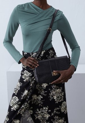 Femme portant un haut à manches longues turquoise et une jupe noire à motifs floraux, tenant un sac à bandoulière noir texturé avec une fermeture dorée.