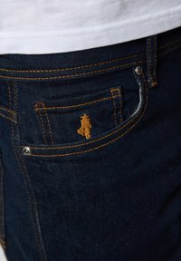 Tamno plave traperice s narančastim šavovima, s malim izvezenim detaljem na džepu i metalnom zakovicom sa strane.